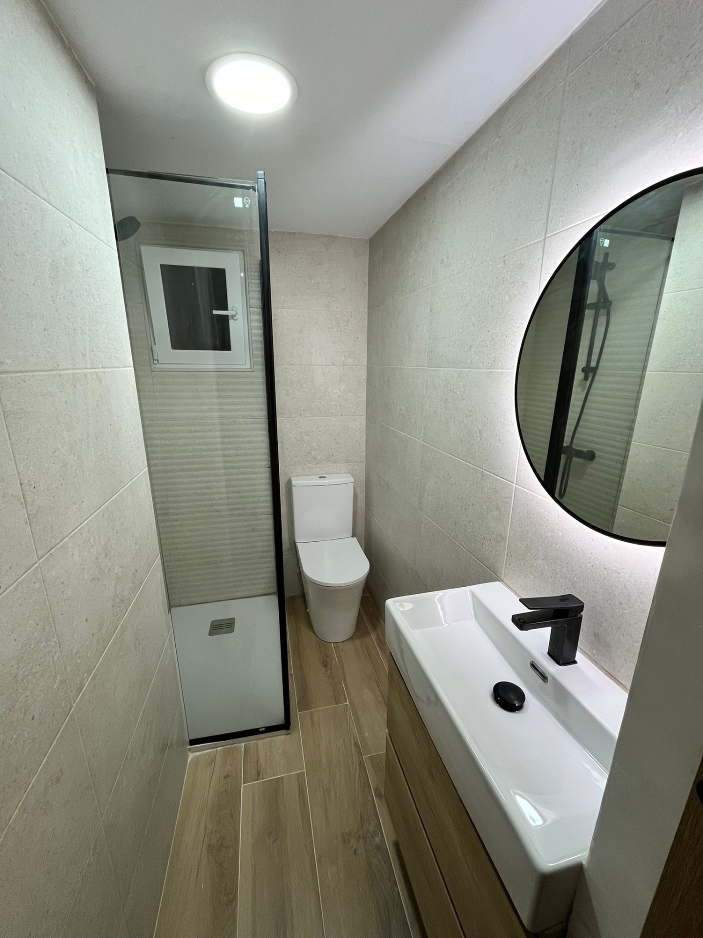 Baño estrecho reformado con plato de ducha, mampara negra y espejo circular retroiluminado